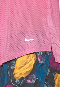 Růžový sportovní top Nike z prodyšného materiálu s krátkými rukávy, s bílým logem a bočními rozparky, v kombinaci s barevnými šortkami s květinovým vzorem.