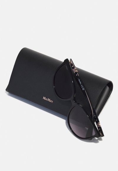 Occhiali da sole neri con lenti scure, forma oversize a occhio di gatto e tempie accentuate in oro, posizionati su un caso in pelle nera con logo impresso.