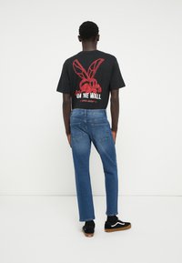T-shirt gráfica preta com texto vermelho "ON THE WALL" e um design de cartoon nas costas, combinada com jeans denim reto azuis e ténis pretos.