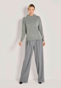 Frau, die einen grauen Langarm-Pullover und weite graue Hosen trägt, steht vor einem schlichten hellen Hintergrund.