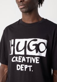 Black cotton t-shirt featuring bold white graphic text: "HUGO CREATIVE DEPT." Relaxed fit with crew neckline and short sleeves.
