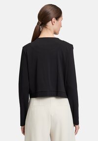 Kobieta z ciemnobrązowym kucykiem, ubrana w czarną, długą bluzkę z krótkim przodem i białe spodnie z wysokim stanem, widziana z tyłu.