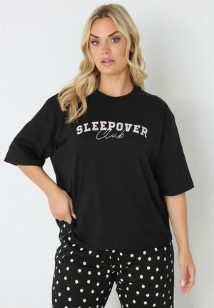 Mujer con cabello rubio largo lleva una camiseta negra que dice "Sleepover Club" y pantalones negros con lunares blancos sobre un fondo liso.