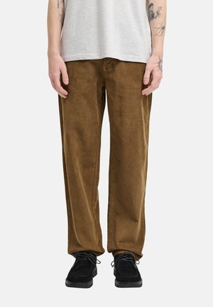 Osoba ubrana w brązowe spodnie sztruksowe, czarne sznurowane buty i jasnoszary t-shirt, stojąca z rękami luźno opuszczonymi wzdłuż ciała.