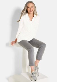 Weißes Langarmshirt mit Knopfleiste und vertikalen Falten, kombiniert mit grauen, cropped Jeans und grauen Sneakers, sitzend auf einem weißen Podest.