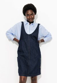 Robe salopette en denim bleu foncé avec poches latérales et bretelles ajustables, superposée sur une chemise rayée bleu clair. Texture de tissu lisse.