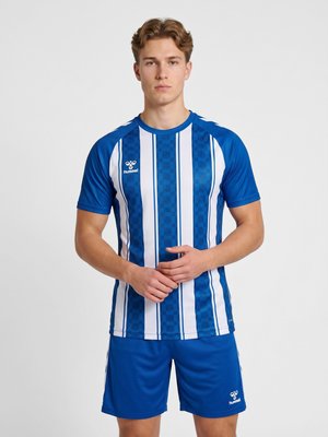 Jonge man in een blauw-wit gestreept sportshirt en blauwe korte broek staat met gevouwen handen.