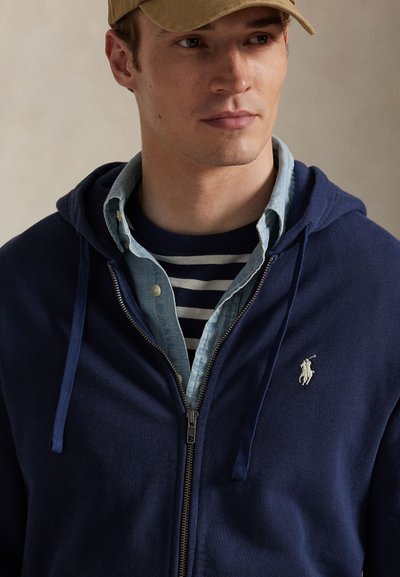 Tmavomodrá mikina s kapucňou na zips vyrobená z mäkkej látky, so zapínaním na zips vpredu, klokanie vrecko a malým vyšitým logom na ľavej strane hrudníka.