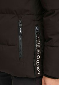 Čierna zateplená bunda s vreckami na zips, s logom značky pozdĺž strany. Hladká tkanina s mierne lesklým povrchom.