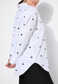 Witte overhemd met zwarte hartpatronen, lange mouwen en een gebogen zoom. Gemaakt van lichtgewicht stof met een gladde textuur.