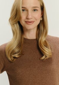 Blonde vrouw met lang golvend haar, draagt gouden creolen en een bruine gebreide trui, glimlacht lichtjes tegen een effen achtergrond.