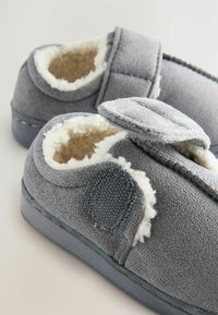 Chaussons en tissu gris avec doublure blanche en peluche, fermeture à bande auto-adhésive, forme arrondie et semelles plates. Texture douce avec des détails cousus.