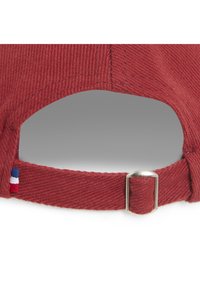 Casquette de baseball en velours côtelé rouge avec une sangle en tissu réglable et une boucle en métal. Elle présente une petite étiquette bleu, blanc et rouge sur la sangle.