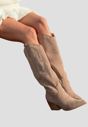 Jambes portant des bottes de cowboy en daim beige montant jusqu'au genou, avec des broderies décoratives et un talon bloc en bois, associées à une jupe blanche à volants.