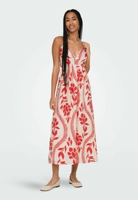 Robe maxi en tissu léger avec un motif floral rouge, décolleté en V profond et bretelles ajustables. Portée avec des ballerines beiges, affichant une silhouette fluide.