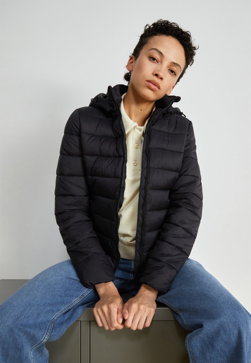 Chaqueta acolchada negra con capucha, que presenta secciones acolchadas horizontales, cremallera frontal y un ajuste casual. Lleva puesta una camiseta polo beige.