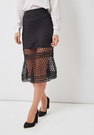 Falda lápiz negra con un diseño en capas que presenta un patrón de punto texturizado. Cintura elástica y sección inferior transparente, combinada con tacones negros.