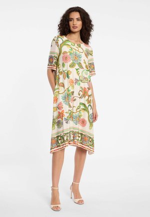 Vrouw met krullend haar, gekleed in een crèmekleurige jurk tot op de knie met levendige bloemen- en kronkelende wijnrankpatronen, en beige sandalen met hoge hakken.