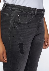 Black distressed denim jeans featuring scattered black studs, a front pocket, and a frayed tear on the thigh area. Slim fit.