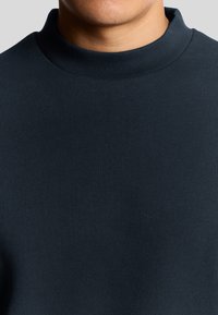 Donkerblauwe trui met een hoge, ribbelige kraag. De stof lijkt glad en gestructureerd, en past snug tegen het lichaam met een minimalistisch ontwerp.