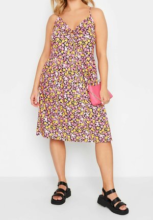 Femme portant une robe sans manches à fleurs roses, jaunes et blanches, tenant une pochette rose et des sandales compensées noires.
