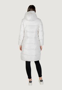 Cappotto imbottito bianco di lunghezza media con cappuccio, caratterizzato da un design trapuntato e una texture liscia. Indossato sopra leggings neri e scarpe bianche.