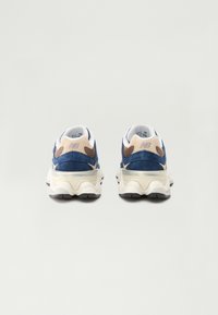 Blue suede sneakers with beige accents, a cushioned sole, and a textured back. Features a white collar and a reflective silver detail.