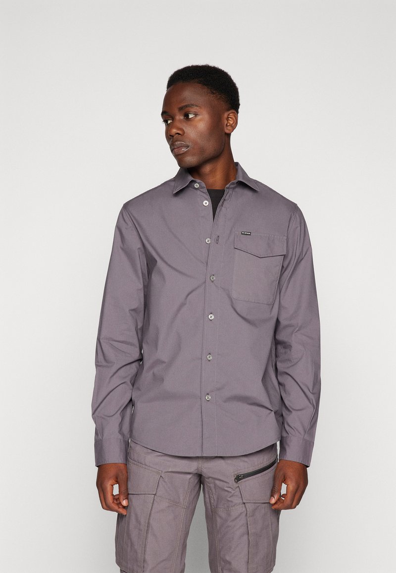 Camisa de manga larga, abotonada, en tejido gris claro con un bolsillo en el pecho, cuello y botones plateados; combinada con pantalones cortos cargo grises.