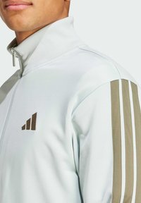 Ljust blå zip-jacka i slät tyg med ståkrage, brun logotyp och olivgröna samt vita ränder på axeln.