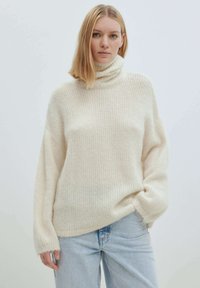 Hellbeige Rollkragenpullover aus weichem, geripptem Material mit lockerer Passform und überschnittenen Schultern. Getragen mit hellblauen Jeans.