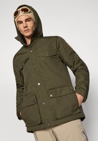 Giacca parka isolata verde oliva con chiusura a zip e bottoni, due tasche sul petto, due tasche inferiori e cappuccio staccabile.