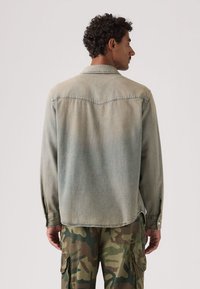 Άνδρας με σγουρά μαλλιά που φοράει φθαρμένο τζιν μπουφάν και πράσινο παντελόνι παραλλαγής cargo, με την πλάτη του προς τα εμπρός σε απλό φόντο.