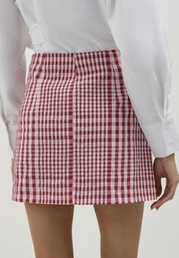 Jupe courte à carreaux rouges et blancs avec des plis, fabriquée en tissu léger. Elle présente une taille lisse et un ourlet qui se trouve au-dessus de la mi-cuisse.