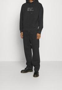 Felpa nera con cappuccio e tasca frontale, testo del logo bianco. Abbinata a pantaloni neri a vestibilità ampia e scarpe nere con dettagli di cucitura gialla.