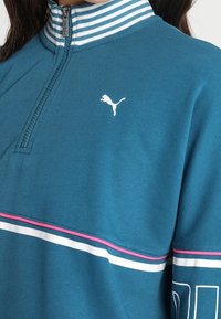 Tealpumpad zip-tröja med randig krage, med en vit Puma-logotyp och horisontella rosa och vita ränder över bröstet.