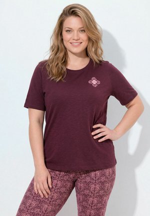 Lachende vrouw met golvend blond haar draagt een kastanjebruin T-shirt en patroon mauve leggings, staand met één hand op haar heup.