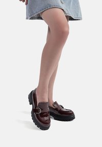 Mocassini patinati bordeaux con suole nere spesse, caratterizzati da dettagli hardware decorati e una finitura liscia e lucida.