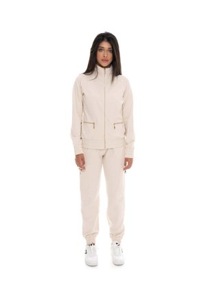 Une femme porte une veste zippée couleur crème et un pantalon de jogging assorti, avec des baskets blanches, sur un fond blanc uni.