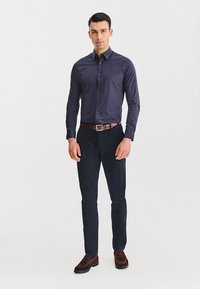 Mężczyzna w ciemnoniebieskiej koszuli w wzory, granatowych spodniach, brązowym pasku i brązowych loafersach, stojący na tle białym.