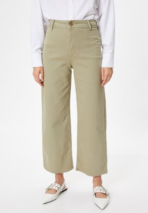 Pantalons larges vert clair à taille haute, en tissu texturé, avec une fermeture à boutons à l'avant et un ourlet effiloché, associés à des chaussures blanches à bout pointu.