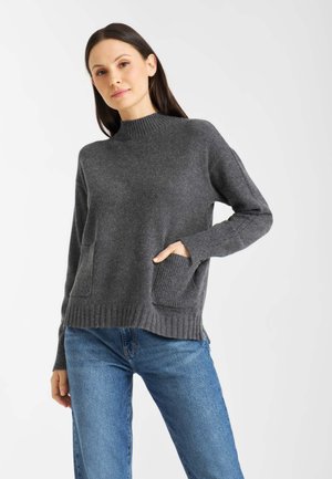 Donkergrijze gebreide trui met een hoge hals, geribde manchetten en twee voorkruisen. Gelaagd stof en een relaxte pasvorm. Gecombineerd met blauwe jeans.