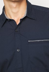 Námořnická košile na knoflíky s vertikálními pruhy, šedým límcem, levým kapsičkovým prostorem a černými knoflíky. Vzhled látky vypadá měkce.