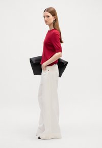 Červený krátký rukáv pletený top v kombinaci s bílými širokými kalhotami. Modelka drží černou koženou tašku. Jednoduchý design, hladká textura a kontrastní barvy.