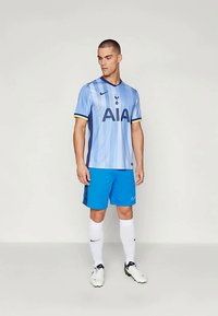 Maillot de football bleu clair avec des accents et des rayures navy, associé à un short bleu vif. Des chaussettes blanches et des crampons noirs complètent la tenue.