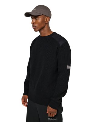 Mann trägt schwarzen Strickpullover mit Schulterpatches und schwarze Hose, schaut zur Seite, trägt auch eine graue Mütze und goldenen Creolen-Ohrring.