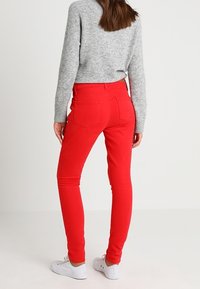Jeans rojos ajustados con una textura suave, bolsillos traseros y una cintura baja, combinados con un suéter gris y zapatillas blancas.