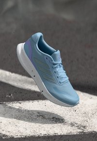 Hellblauer Sportschuh mit Mesh-Obermaterial, flexiblem Sohlenprofil und drei grauen Streifen. Verfügt über einen violetten Akzent an der Seite und ein strukturiertes Design.