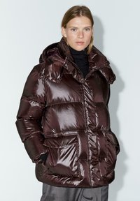 Donna che indossa un giubbotto imbottito lucido di color marrone scuro con cappuccio, dolcevita nera e pantaloni grigi, in piedi contro uno sfondo chiaro e semplice.