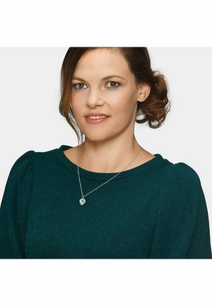 Silberne Halskette mit einem runden Anhänger, der einen hellblauen Edelstein zeigt, getragen über einem strukturierten türkisfarbenen Pullover mit puffartigen Ärmeln.