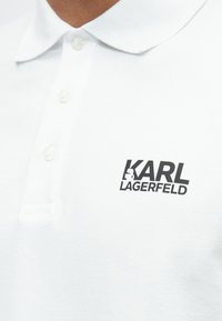 Fehér, pamutból készült galléros póló, bal mellrészen fekete "KARL LAGERFELD" logóval, klasszikus gallérral és három gombbal.